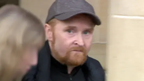 Picture of Scott Forbes near the High Court in Glasgow. He has ginger hair and shortly chopped beard and he is looking towards the camera. Forbes is wearing a gray tweed style baseball cap. 