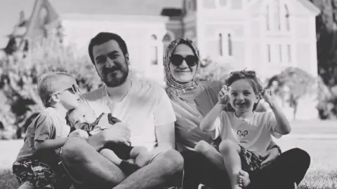 Instagram Zehra Gultekin and her husband Bilal Gultekin smile in a black and white photo while sitting with their children on a lawn in front of a large building. Zahra is wearing a headscarf and sunglasses, and a child – on the far left of the photo – is also wearing sunglasses