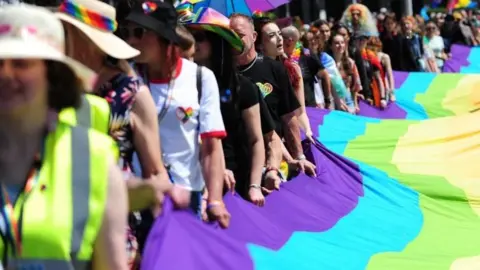 A line of people carrying a large pride flag. It is cropped so the colours of the rainbow include purple, blue, green, yellow, orange. There are people in brightly coloured clothing carrying the flag. 