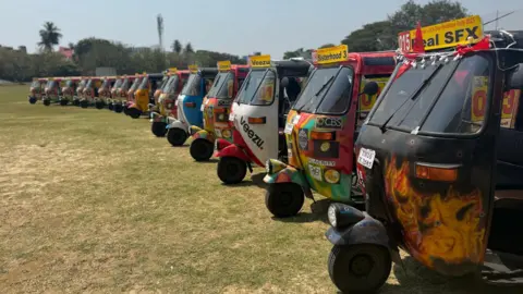 Claire Jenkins A row of about 17 colourfully decorated and painted tuk-tuks on a mown lawn in India. There are trees in the background under a bright blue sky.