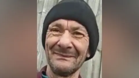 Metropolitan Police Portrait of Gheorghe Trica smiling and wearing a black beanie hat against a wooden fence background.