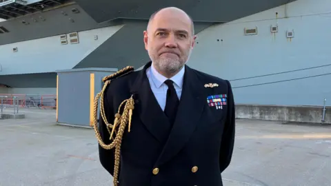 Vice Adm Stephen Moorhouse, CBE stands on Portsmouth Naval Base in front of HMS Prince of Wales. He is wearing a Royal Navy uniform consisting of a darkj blue blazer, with a golden cord trailing down from his left shoulder into his breast pocket. He is bald with short grey stubble.
