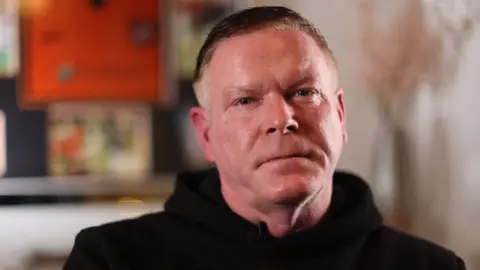 A 53-year-old man with short brown hair and a black hooded top looks at the camera