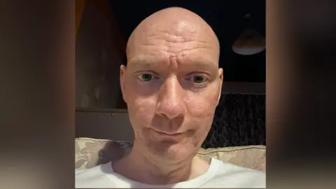 Raymond Osborn A selfie-style image of Raymond Osborn. He is smiling at the camera and he sits on a cream coloured sofa. He is bald and wears a white top. 