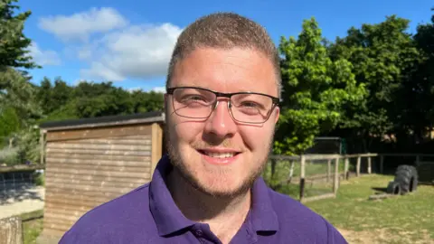 Tom Kitcher had short-cropped fair hair and is wearing black-rimmed glasses and a purple polo-neck jumper.