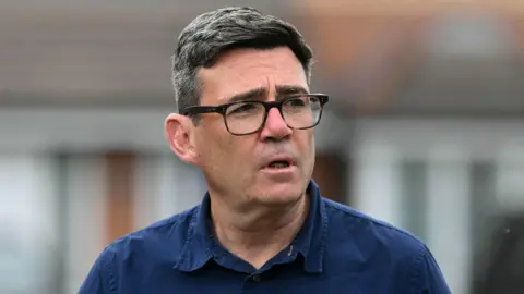 Andy Burnham photographed from the shoulders up. He is wearing a navy blue shirt and black glasses. He is not looking at the camera.