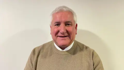 Councillor Patrick Harley. He has short white hair, brown eyes and is clean shaven. He wearing a brown jumper and is stood in front of an off-white background. 