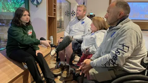 BBC Young Reporter Marianna is sitting down and holding a microphone as she interviews a woman and two men who do curling training at the Flower Bowl. The three players, all looking at Marianna , are in wheelchairs and wearing grey tops with The Flower Bowl printed on them. Marianna is wearing a green England Curling Association hoodie, with the ECA logo featuring the English flag. 