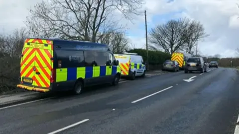 Along the left-hand side of the road, several police vehicles are parked on the verge and roadside. Closest to the camera is a large dark blue police van with high-visibility yellow and blue markings. The rear doors feature bright fluorescent red and yellow chevron patterns designed to improve visibility. Further ahead, another marked police van is parked behind it. In the distance, a third emergency vehicle displaying similar chevron markings is also visible.