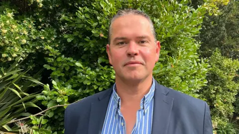 Marcus Calvani is standing in a garden smiling at the camera. He is wearing a blue and white striped shirt under a navy jacket. He has short grey hair combed back from his face.