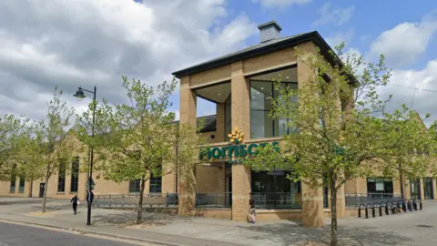 The Morrisons supermarket is a large, modern brick building. The name is in large letters across the front, partially covered by branches from trees planted on the pavement in front