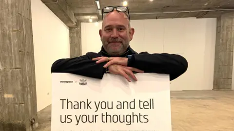 BBC A man in a dark top stands behind a sign saying "thank you and tell us your thoughts"