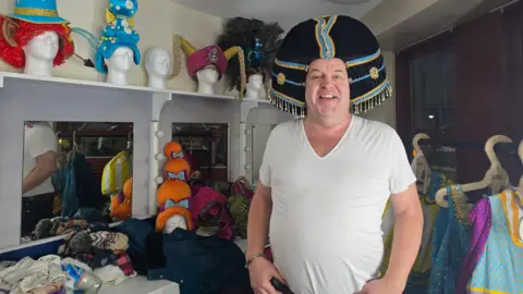 Hayley Coyle/BBC A man in a white t-shirt stands in a dressing room with a black wig on his head, with gold and blue bands and braids around it. It resembles Cleopatra's hair. Behind him, a clothes rail with brightly patterned costumes hanging on it. On the other side of him are three mirrors on a wall, a shelf above them with mannequin heads with different colourful wigs and headpieces on them.