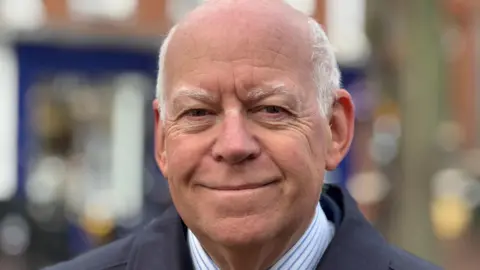 West Berkshire Council An older man is smiling at the camera, wearing a suit and tie. It looks like a shop is blurred behind him.