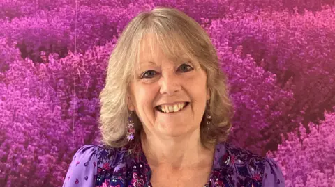 A woman wearing a purple dress and earings, with shoulder length hair, smiling at the camera. She is stood in front of a backdrop of purple flowers.