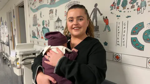 Natasha, wearing a black jumper, is holding her baby. The baby is wearing a maroon hat and baby-gro. They are in a hospital corridor.