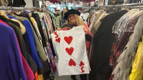 Brian Farmer/BBC A woman wearing a black hat and glasses holds a top which looks like a three of hearts playing card. She is standing next to racks of clothes