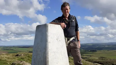 Patrick Norris Patrick Norris is standing by a trig point with rolling hills behind him. He is a middle-aged man with a weather-beaten face wearing brown trousers and a blue top 