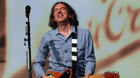 Gary Lightbody, the singer of Snow Patrol, onstage playing a guitar with a microphone in front of him.  He has dark hair to his shoulders and is wearing a blue shirt. 