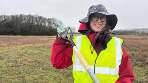 Tricia wears a yellow high-vis vest, a red raincoat, a black rain hat, gloves and round glasses. She is smiling at the camera and holding a shovel by the handle up at the camera. 