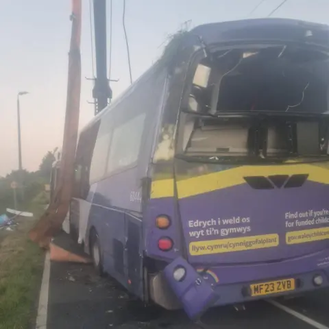 Jacko Thomas A view of the purple damaged bus after the crash from the back. The back window is smashed, with a light panel hanging loose. A long band hangs down the side of the bus with a piece of machinery in the background. 