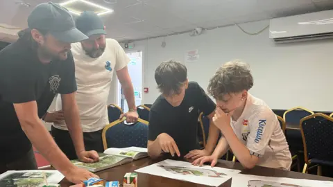 Owen Sennitt/BBC Two men wearing hats and two boys look at a table with printouts of skatepark designs inside a social club