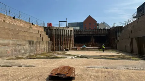 Andrew Spence / LDRS View of the inside of the dry dock a large rectangular concrete structure with a dock gate in the corner and the tops of buildings in the distance