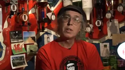 A screenshot of a man in a red top with a circular logo on it. He is wearing a black cap. He has grey hair just above his shoulders and has large thin-rimmed glasses. Behind him is a red wall and several bottles and posters are hanging up.