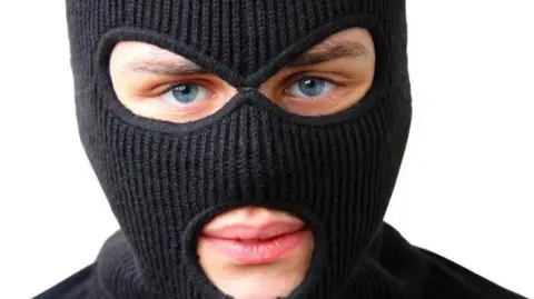 A close-up of a man wearing a black, full-face balaclava. He is standing in front of a white background.