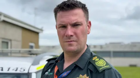Will Bellamy, who has short dark hair and is wearing a green ambulance service uniform.