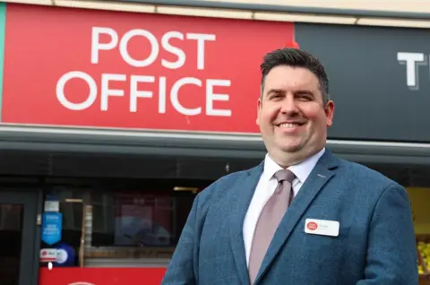 A man with short dark hair and a blue jacket with a brown tie and Post Office name badge is standing in front of a large red Post Office sign on a building.