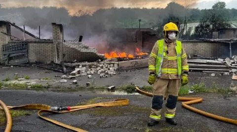 Shows a firefighter wearing a yellow hat and facemask yellow gloves and firefighting gear standing in front of a burning building.