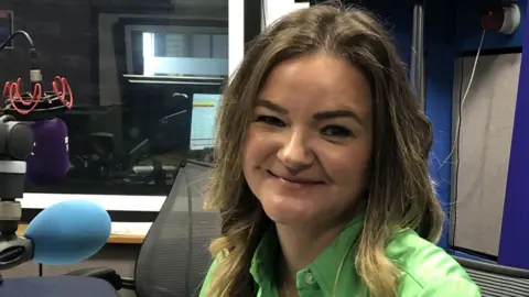 A woman is sitting in the radio studio at BBC Radio Suffolk. She has long hair and is wearing a bright green shirt.