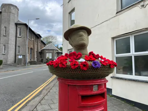 A post box in Criccieth at Y Neuadd Coffa with a basket on the top filled with red flowers and the bust of a soldier in a helmet in grey.