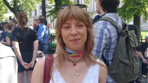 A woman with blonde hair, looking at the camera and smiling. She is wearing a white top, necklaces and sunglasses on her head. There is a crowd of people behind her.