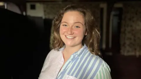 BBC Alexandra Godfrey smiling at the camera. She is a young woman with a blue and pink striped shirt on and has curly brown hair.  The background is dark because she is at a silent disco. 