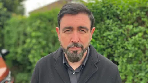 Doherty has dark hair and a beard speckled with grey. He is standing in front of a hedge. 