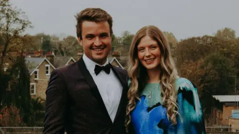 Jack Schofield A young couple stand side-by-side smiling. On the left, the man wears a black suit with a white shirt and black bow tie. He has brown hair and brown stubble. On the right, the woman has long curled blonde hair and wears a dress with blue and green patterns.