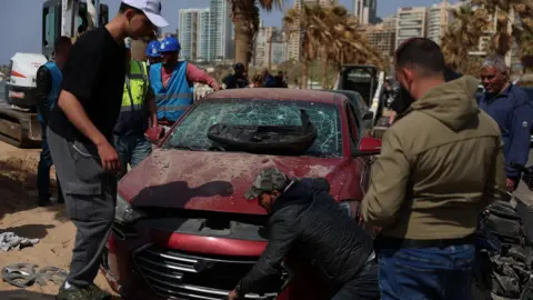 Lebanese officials and civilians inspect a car damaged in an Israeli strike on Ramlet al-Baida beach in Beirut, Lebanon (12 March 2026)