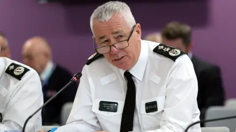 PA Media PSNI Chief Constable Jon Boutcher, sitting at a table with a microphone in front of him. He has short grey hair and is wearing glasses, a white shirt and black tie. 