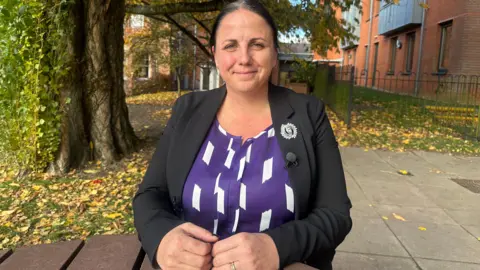 Vikki Rundle-Brown, who has dark hair tied back, wearing a purple top and black jacket sat on a wooden picnic bench in front of grass and a cement paved area.
