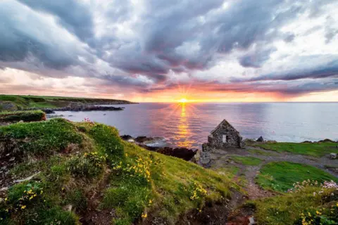Sam Ross A coastal sunset with part of a ruined house on a clifftop, and the sun on the horizon under a pastel sky.