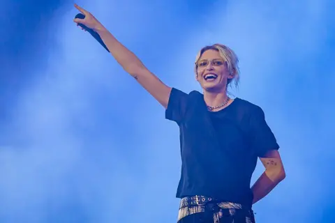 Getty Images Emily Armstrong performs on stage and holds her hands up. She is wearing a black t-shirt and black belt. She has blonde hair tied up, she is wearing yellow glasses. 