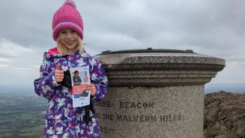 A young girl with blonde shoulder-length hair smiles as she stands at the top of the Worcestershire Beacon, next to the marker at the top, holding an NHS leaflet with information on about plasma donation. She is giving a thumbs up with her other hand and is wearing a pink beanie hat and a purple coat with unicorns on.