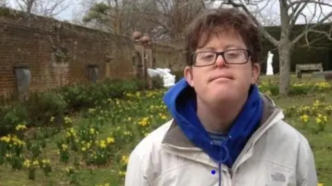 A man wearing a white coat, blue hoodie and glasses stood in a garden with yellow flowers.