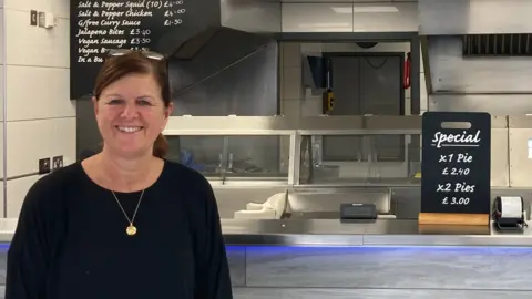 Kim Ashplant wearing a black top, gold necklace. Her hair is tied up. She is stood in front of the chip shop counter and fryers. 