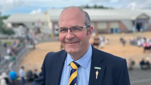 A man stands in front of a show ring at the Devon County Show. There are horses in the ring in the background. He is wearing a blue shirt and yellow tie under a dark blue blazer with two small badges on the lapel.