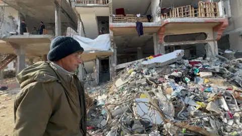 Nabil in profile, wearing an olive-coloured overcoat, glasses and a dark green woolly hat, stands by a big pile of rubble 