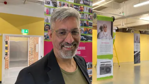 A silver-haired man man wearing glasses who is smiling while standing in front of a couple of posters hanging from a ceiling.