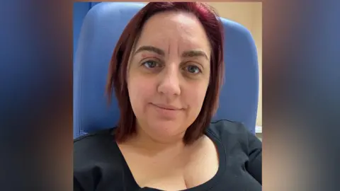 Zara Dedman A woman with dark red hair smiling at the camera. She is wearing a black top and is sat on a blue chair. 